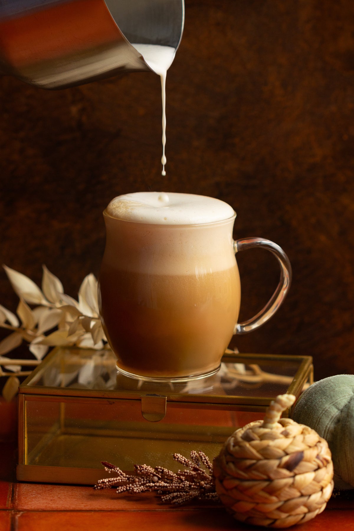 Cozy aesthetic scene of milk dripping into a very full chai latte in a glass mug. Scene is set with braided grass pumpkins, dried flowers, and moody lighting.