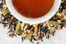 Close up of dry and brewed Pantry Hill Blend. The dry tea is composed of black tea leaves, ginger, crushed cardamom pods, and cinnamon around a white cup with dark reddish-brown brewed tea.