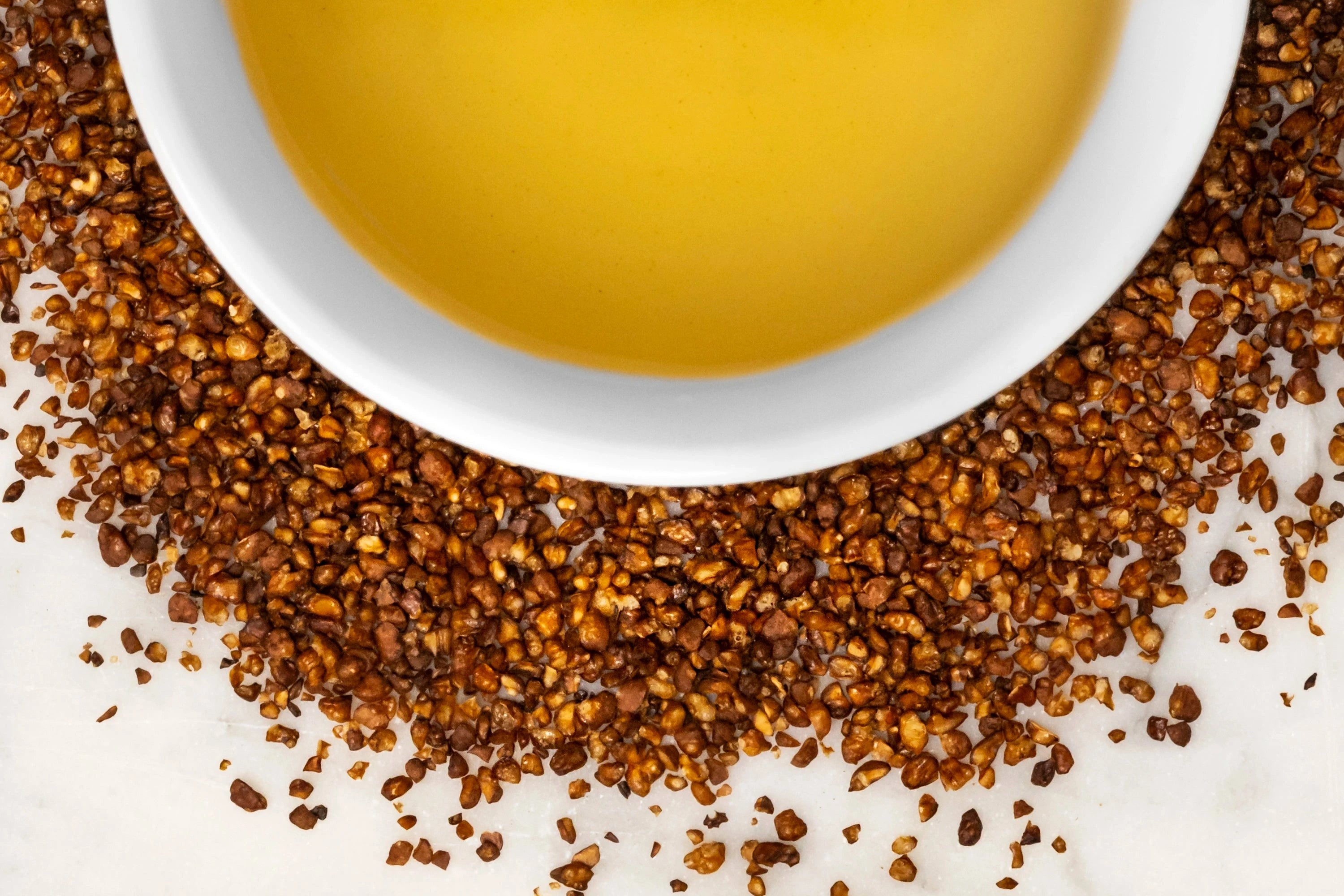 Close up of dry and brewed Sobacha. The dry, round grains of roasted Sobacha are displayed around a white cup filled with golden-hued brewed tea.